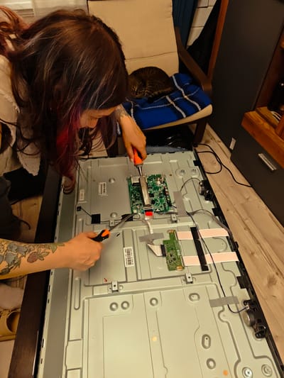 A person with long, dark hair with red and purple streaks is shown from the chest up, bent over a disassembled television set on a wooden floor. The person is wearing a grey top and has a tattoo of flowers and leaves on their left arm. They are holding two screwdrivers, one orange and one black and orange, and appear to be working on the electronic components of the television. The television's back panel is removed, revealing circuit boards and wiring. In the background, a cat is sleeping on a chair with a blue and white striped blanket. The environment appears to be a home. The exact location within the city of Свистуха (кузяевский С/о), Russia cannot be determined from the image alone, but the scene depicts an indoor domestic setting.