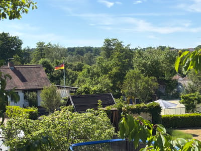 View from an elevated position overlooking a residential area in Zirndorf, Germany. The German flag is prominently displayed in the middle ground. A variety of houses are visible, including one with a red tile roof on the left. Dense foliage fills the landscape with mature trees and bushes. In the foreground, there are leaves of a tree. A trampoline is visible in the lower portion of the view. In the midground, a black building and greenhouse are present. The sky is light blue with some scattered clouds.