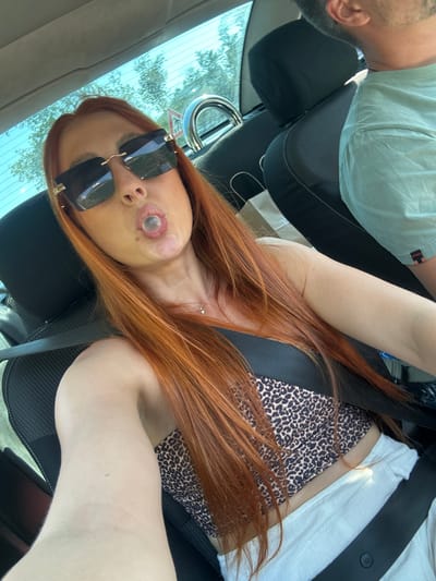 The image shows two people in a car. The person in the foreground is a woman with long, auburn hair and sunglasses. She is wearing a leopard print top. She appears to be taking a selfie, as the camera is angled towards her. Her facial features are distorted due to the angle and a large, circular object is obscuring part of her face, possibly a bandage or a piece of equipment. She has a slight smile, but the overall expression is difficult to discern.
The second person, partially visible in the background, is a man wearing a light green t-shirt. He is seated in the driver's seat, and his profile is visible.
The scene is set inside a car, with car seats and seatbelts visible. The background shows trees and foliage through the car window, suggesting an outdoor setting, likely during the daytime given the lighting. The weather appears to be clear. There are no discernible emotions conveyed by the man. The image is taken in Vrijesno, Montenegro.