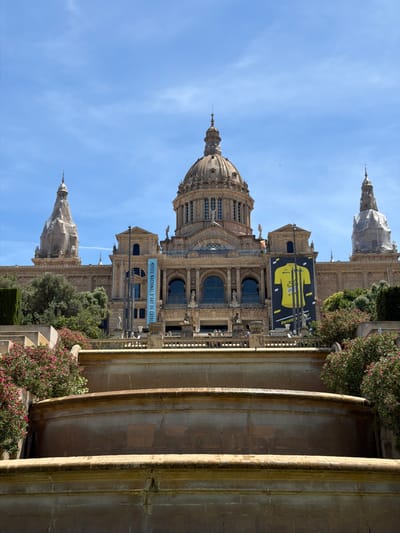 The image shows the Palau Nacional, also known as the Museu Nacional d'Art de Catalunya, located in Barcelona, Spain, on Montjuïc hill. The building features a large central dome and two smaller domes on either side, with ornate architecture and stone detailing. In front of the building, a large banner displaying a yellow cycling jersey with text that reads "TOUR DE FRANCE" is visible, indicating an event related to the cycling race. Another blue banner with text that appears to be in Catalan, "MUSEU NACIONAL D'ART DE CATALUNYA," is also present. Below the building, there are tiered fountains and lush greenery, including blooming pink flowers. The sky is clear blue with a few wispy clouds. There are no individuals clearly depicted in the foreground or engaged in any specific actions. The image captures a grand architectural landmark under a bright, sunny sky.