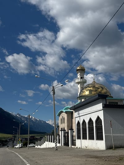 The image shows a mosque with a golden dome and minaret situated in the city of Verkhnyaya Teberda, Russia. The mosque is a white building with arched windows featuring black metal grilles. In the background, a road curves into the distance, lined with streetlights and leading towards a town nestled at the foot of snow-capped mountains. The sky is a vibrant blue with scattered white clouds, and a power line runs diagonally across the upper portion of the frame. Several cars are visible on the road, and some white fences line the side of the street. The overall scene depicts a serene, mountainous landscape with a prominent religious structure.