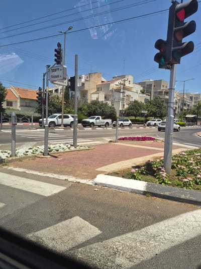 The image captures a street view in Kfar Saba, Israel, on a clear, sunny day. The scene focuses on a pedestrian crossing at an intersection. A traffic light, displaying a red signal for vehicles and a green walking person symbol for pedestrians, is prominently visible on the right. A street sign in Hebrew and English reads "Weizmann st." on the left side of the intersection. Several vehicles are present, including white SUVs and a pickup truck, either stopped at the intersection or driving through. The environment includes buildings in the background, streetlights, power lines, and a median strip with flowering plants. The perspective appears to be from inside a vehicle, looking out through a window.