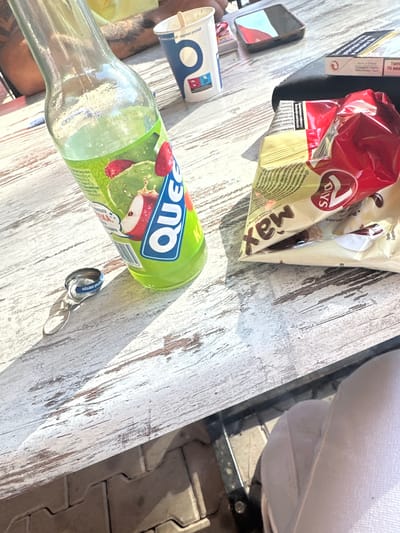 A table with a bottle of "Quez" juice, a bag of "7 Days Max" croissants, a cigarette pack, a smartphone, a cup, and a bottle cap is captured from a high angle. The table is made of wood and has a worn-out texture with white paint and visible wood grain. The scene is set outdoors on a sunny day, with shadows cast on the table, suggesting a possible location in Cherven Bryag, Bulgaria.