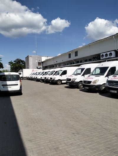 In Șoldănești, Moldova, a fleet of white commercial vans is parked in a line on a paved lot in front of a long, white industrial building under a clear blue sky with scattered clouds. The vans, several of which appear to be Mercedes-Benz Sprinters, are arranged in a row, facing towards the right of the frame. Some vans have logos visible, including "Rudetel" on a few of them. The building has large windows and several industrial air conditioning units mounted on its facade. In the foreground, a smaller white Dacia Dokker van is parked at an angle, facing left. The ground is paved with interlocking grey bricks laid in a herringbone pattern. A shadow is cast diagonally across the foreground from the left side of the image. There are no people visible in the scene.