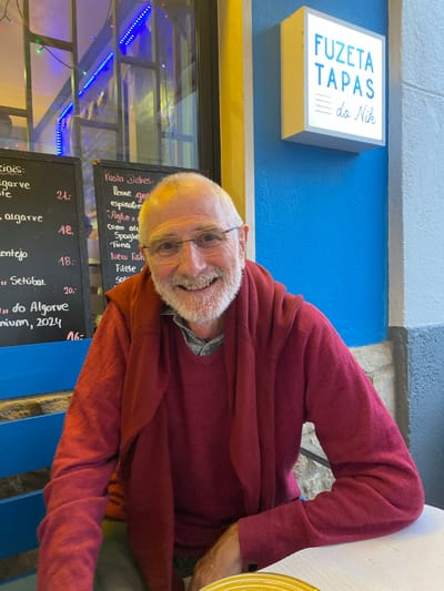The image shows a man with white hair and a beard, wearing glasses and a red sweater, sitting at a table. He is smiling and looking towards the camera. Behind him, there are two chalkboards with handwritten menus. The chalkboard on the left lists "algarve" and "alagarve", with prices. The chalkboard on the right lists "Pasta Dishes", "Penne gen", "espinagre", "Agilo e", "corn ai", "Spaghe", "Tuna", "New Fish", "Filete", and "Sole", with prices. To the right of the man, a illuminated sign reads "FUZETA TAPAS do Nik". The background also shows a blue wall and part of a window with blue and green fairy lights. The scene appears to be an outdoor seating area of a restaurant in Olhão, Portugal.