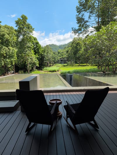 A serene outdoor seating area overlooks a lush, green landscape with a water feature and distant mountains under a partially cloudy blue sky. In the foreground, two dark-colored lounge chairs are positioned on a dark wooden deck. A small, wooden side table with a carved top sits between the chairs. The chairs face a rectangular water feature that appears to be a shallow pool or reflection pond, with a section of it designed as a waterfall. Beyond the water feature, a manicured lawn stretches towards a cluster of modern buildings nestled amongst trees. The background is dominated by a dense forest of green trees, with rolling hills and mountains rising in the distance. The overall impression is one of tranquility and natural beauty. The scene is likely located in Ban Pong Nuea, Thailand, given the tropical vegetation and architectural style. There are no people visible in the image.