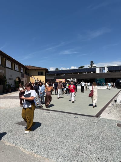 In Florence, Italy, on a sunny day, a crowd of people are gathered in an outdoor plaza. The plaza is paved with small, light-colored stones. Buildings with terra cotta roofs and tan facades line one side of the plaza, with signs visible for "FLY3," "DISTRstressed," and "ANGELO." Another building on the opposite side of the plaza has a darker, modern facade with large windows and banners displaying the text "FANTASTIC CLASSIC."
In the foreground, a woman with brown hair is walking away from the camera, wearing a white top and olive green, textured pants, and carrying a black folder. She is looking down at a mobile phone in her hands.
To her right, a group of people are walking and talking. One man is wearing a light blue blazer and white pants, while another is wearing a red shirt and white pants, holding a yellow ball. A woman with dark hair is walking away from the camera, wearing a patterned dress and carrying a large red tote bag.
In the background, more people are seen mingling, and a black, cylindrical object, possibly a camera or speaker, is positioned at the edge of the frame. The sky is a clear, bright blue with a few wispy white clouds. The overall scene suggests a public event or gathering in an urban setting.