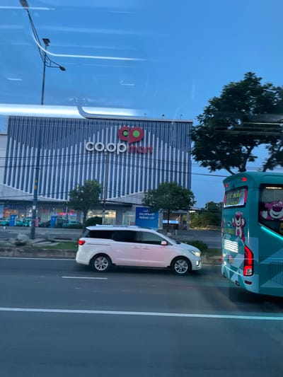 A white minivan is driving on a road in front of a Co.opmart store in Thuận An, Vietnam. A blue bus is driving on the right side of the road next to the minivan. The Co.opmart store has a blue and white striped facade with the Co.opmart logo at the top. There are trees and streetlights visible in the background. The sky is a dusky blue, suggesting it is either early morning or late evening.