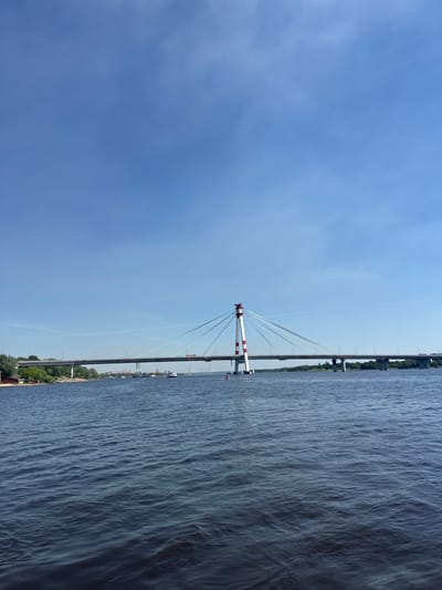 The image shows a cable-stayed bridge spanning a wide river under a clear blue sky. The bridge features a distinctive red and white striped tower supporting the cables. In the foreground, the dark blue water of the river is visible with moderate ripples. In the background, the far bank of the river is lined with trees and buildings, suggesting an urban or semi-urban environment. A few boats are present on the water, and vehicles can be seen on the bridge itself. A red buoy floats near the bridge tower. The overall scene depicts a sunny day with a focus on the bridge and its relationship with the waterway. This scene is likely the Severny Bridge over the Yagorba River in Cherepovets, Russia, given the distinctive bridge design and the geographical context of the Yagorba River flowing through the city. There are no people directly visible in the frame.
