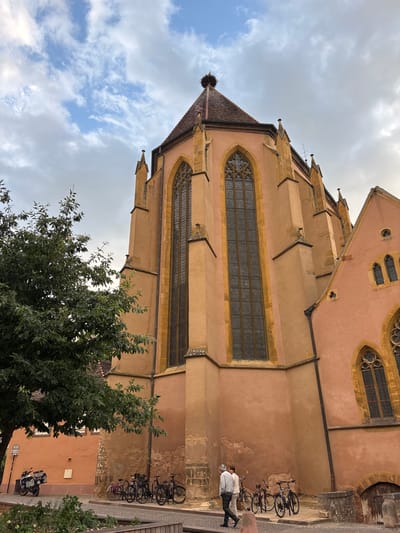 The image shows the Saint-Martin's Church, also known as the Collegiate Church of Saint-Martin, in Colmar, France. The church, with its characteristic salmon-pink facade, features tall, arched windows with intricate tracery. A prominent detail is a stork's nest atop the tiled roof. In the foreground, a tree with lush green foliage partially obscures the church. To the left, a motorcycle is parked on the cobblestone street. Several bicycles are leaned against the church wall. Two individuals, a man and a woman, are walking away from the camera on the paved pathway, with the woman appearing to be wearing a light-colored top and dark trousers, and the man in a light-colored top and dark trousers. The sky above is partly cloudy, with a mix of white and gray clouds against a blue background. The street is paved with cobblestones, and there is a low stone wall and some decorative planting beds. The overall scene depicts a quiet street in a historic European city.