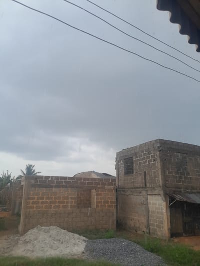 The image depicts an outdoor scene in Ifo, Nigeria, under a heavily overcast, grey sky with visible rainfall. In the foreground, three parallel electrical wires traverse the upper portion of the frame, with a segment of a brown corrugated roof visible in the top right corner. The ground level features two piles of construction materials: a light grey mound, consistent with sand or cement, on the left, and a darker grey pile of aggregate (gravel/crushed stone) on the right. Sparse green vegetation grows between these piles and alongside the structures.
Two primary buildings are visible in the mid-ground. To the left is a single-story structure constructed from orange-brown clay bricks, appearing incomplete with open wall sections and no roof. To its right stands a taller, two-story building made of grey concrete blocks, also appearing unfinished or in a state of disrepair, featuring open window apertures and an exposed concrete slab for its first floor roof. A small, roughly constructed shelter with a corrugated metal roof is attached to the lower right side of this grey block building. In the background, beyond these structures, additional obscured buildings and vegetation, including a palm tree, are partially visible. No individuals or active interactions are present within the frame.