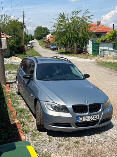 The image depicts a silver BMW 3 Series Touring (E91 generation) parked on a gravel and dirt patch in a rural or semi-rural setting. The car is viewed from a front-right angle, with its front grille, headlights, and roof rails clearly visible. The iconic BMW logo is prominently displayed on the front grille. A Bulgarian license plate, "EH 2066 KX," with the "BG" country code on the blue EU strip, is attached to the front bumper.
The car is situated alongside a narrow, unpaved road that stretches into the distance. The surrounding environment features several houses, including one to the left with an orange-tiled roof and green accents, partly obscured by a large, leafy tree. Further down the road, utility poles with power lines are visible, along with another parked vehicle, which appears to be a dark SUV or truck. The ground immediately surrounding the BMW is a mix of gravel, dirt, and patches of grass, bordered by a low brick structure towards the bottom right.
The scene is captured during bright daylight, indicated by strong shadows, suggesting midday or early afternoon on a clear, sunny day with some scattered white clouds in the blue sky. There is no human activity or people visible in the frame; the car is stationary. The overall setting points to a quiet, residential area within a village in Botievo, Bulgaria.