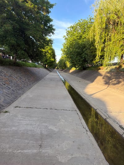 This outdoor image, taken in Târgu Mureș, Romania, presents a linear concrete water canal running diagonally across the scene. The canal, filled with dark, reflective water, separates two distinct ground surfaces. To the left of the canal lies a wide, light grey concrete path with visible cracks and some grass growing through them. Long shadows, possibly from trees and an unseen lamppost (hinted at by its shadow in one crop), stretch across this concrete area, indicating a specific time of day.
To the right of the canal is an embankment. Its lower section features dark, irregular stone paving, while the upper edge is lined with green grass. Beyond this embankment, and across the scene, a dense canopy of lush green trees with abundant foliage dominates the landscape, some appearing lighter green, possibly willows, others darker. Above the trees and concrete structure, a bright blue sky with scattered, wispy white clouds is visible.
No explicit activity is taking place; the scene is static, showcasing the environment. The distinct, elongated shadows and the warm, golden light illuminating parts of the concrete suggest either early morning or late afternoon/evening, commonly known as the golden hour. The clear sky indicates sunny and pleasant weather. While no people are clearly prominent, very small, indistinct figures can be discerned in the far distance under the trees on the left side, hinting at human presence. There is no visible text in the image.