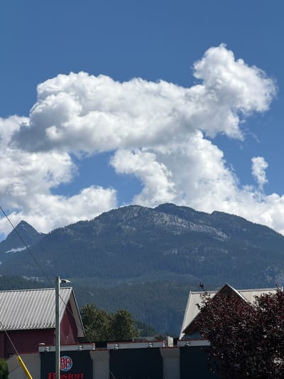 This image depicts a mountain range partially obscured by lush green trees. A vast expanse of the sky is filled with fluffy white clouds against a clear blue backdrop. In the foreground, buildings with corrugated metal roofs are visible. One building features a prominent red section with a white roof, and another has a dark facade with a circular logo that appears to read "BP" and "Boston." Power lines traverse the scene, and a street light is mounted on a pole. The overall setting suggests a semi-urban or commercial area situated near a mountainous natural landscape. The location is in Area D (Elaho/Garibaldi), Canada.