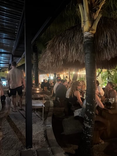 Captured at night in Uluwatu, Indonesia, the image shows an outdoor restaurant scene. A crowd of people is gathered, seated at tables under a thatched roof, suggesting a tropical or coastal environment. Lighting fixtures are positioned above the tables. A palm tree trunk runs vertically across the frame. Pebbles cover the ground. A man wearing a light-colored shirt and dark shorts stands in the foreground.