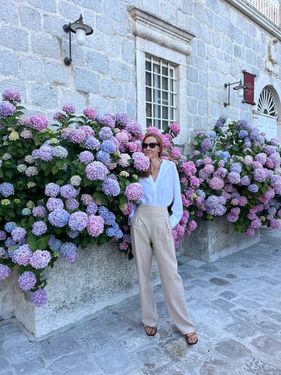 A full-shot image captures a woman standing in front of a stone building adorned with blooming hydrangeas in Perast, Montenegro. The woman, positioned centrally, faces the camera slightly, with her head turned to her left (viewer's right), looking slightly past the lens. She has reddish-brown hair, wears dark sunglasses, a white long-sleeved collared shirt with a V-neck, high-waisted wide-leg beige trousers, and dark brown flat sandals with a toe loop. Her hands are placed in her trouser pockets.
The background consists of a building constructed from large, light grey-blue, rectangular cut stones. On the left side of the building, a tall, narrow window with a white frame and multiple white grid panes is visible. Above this window is a decorative stone lintel. To the right of the window, a black metal wall lamp with a white inverted cup-shaped shade extends from the stone wall. Further right, a red rectangular sign with white text (partially obscured) hangs below another similar wall lamp. On the far right, a white wooden door with an arched fanlight, featuring radiating white spokes, is visible. Above the door, there is a stone arch and a sculpted crest or emblem.
Lining the base of the stone building are dense bushes of hydrangeas, predominantly displaying clusters of pink, purple, and blue flowers, with some lighter green/white new blossoms. These flowers extend from a raised planter or foundation made of the same light grey stone as the building. The ground is paved with irregular, light grey-blue cobblestones or flagstones, showing some patches of darker moisture. The scene is illuminated by natural light, casting soft shadows.