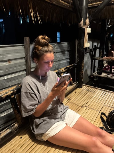 A young woman with her hair in a bun, wearing a grey t-shirt and white shorts, sits on a bamboo mat floor. She is holding a light purple smartphone and appears to be looking at it while wearing white earbuds. The background consists of a corrugated metal wall and a thatched roof supported by bamboo poles. The scene is dimly lit. The location is a communal outdoor seating area, possibly a restaurant or rest stop, in Kertasari, Indonesia.
