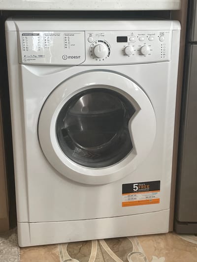A white Indesit front-loading washing machine is centrally positioned in an indoor setting within Ihtiman, Bulgaria, likely a kitchen or utility room. The appliance features a white chassis and a clear, circular front-loading door, behind which the metal drum is visible. The control panel, located on the upper section of the machine, displays various program settings categorized under "EVERYDAY FAST" (programs 1-6 with times ranging from 20 to 59 minutes), "Special & Partial" (programs 7-11), and "Dedicated" (programs 12-16, including specific settings for wool and baby clothes, with temperatures like 60°/40°). An "A+" energy efficiency rating is noted, indicating a capacity of 1-7 kg and a spin speed of 1000 RPM, along with a "Water Balance Plus" feature.

To the right of the program list, a central rotary dial, labeled "My Time," allows selection of the 16 programs; its current position is not definitively set to a specific number. Above the dial, "ON OFF" and "START PAUSE" buttons are visible, alongside a digital display that is currently unlit. Additional control buttons include "Timer," "Extra Wash," and "Energy Saver." Below these, two rotary knobs control temperature (options from 20° to 90°) and spin speed (options from 400 to 1000 RPM). A vertical column of indicator lights with "END" at the bottom suggests program status.

On the lower right facade of the washing machine, an orange and black sticker advertises a "5 Years FREE GUARANTEE ON MOTOR/COMPRESSOR" and lists several customer service phone numbers (800 697 152, 0 800 94 82 20, 800 535 243, 800 235 3434) and a QR code.

The washing machine is situated beneath a light-colored, speckled countertop. To its immediate left is a light brown wooden cabinet or wall. To its right, a dark gray appliance or cabinet is partially visible. The floor beneath consists of beige and brown patterned tiles, with a small portion of a light-colored, textured rug visible on the far left. The appliance appears to be clean and in functional condition. No individuals are present in the image.