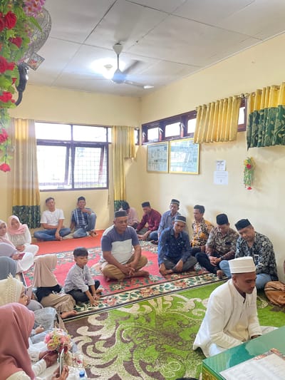 In an indoor room, likely in Pasar Paloh, Indonesia, a group of people are gathered, seated on the floor on a large, ornate rug. The room has light yellow walls and is illuminated by ceiling lights and natural light from windows covered by yellow and green patterned curtains. In the foreground, a man in a white traditional outfit with a white cap is seated on the floor, facing slightly to the right, looking down. To his left, a young boy in a light blue shirt is also seated, looking forward. To the left of the boy, several women are seated, some wearing hijabs in pink and beige. One woman in a white wedding dress with a beaded headpiece is holding a bouquet of flowers and looking at her phone. Further back, a group of men are seated, all wearing traditional attire and many with caps. One man in a white t-shirt and blue jeans is seated cross-legged. A ceiling fan is visible in the upper left corner, along with decorative red flowers. Several framed posters or pictures are on the wall in the background. The overall atmosphere appears to be a formal gathering or ceremony.