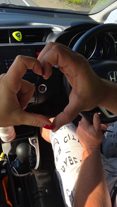 Two people's hands form a heart shape in the foreground. They are inside a vehicle, with the dashboard and steering wheel visible. The person on the left has bright red nail polish. The person on the right is wearing a white t-shirt with a repeating black pattern and a gold bracelet. A black leather wallet is visible on the center console, next to the gear shift. The vehicle appears to be moving, as suggested by the external scenery visible through the windshield. The location is identified as Kruglikovo, Russia.