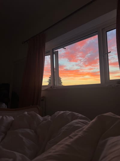 The image depicts an interior view from a bedroom, looking out of a window at a vibrant sunset over the city of Brighton, United Kingdom. The foreground is dominated by the soft, rumpled white duvet of a bed, partially obscuring the lower portion of the scene. To the left, a textured reddish-brown curtain hangs from a curtain rod, partially drawn and casting shadows.
The window itself is a white-framed casement window, with one pane open. Through the open window, the sky is a dramatic spectacle of sunset colors. Fiery oranges and pinks dominate the clouds, transitioning into softer blues and yellows towards the horizon. A silhouette of a tree is visible against the illuminated sky, suggesting an outdoor environment.
The room appears to be dimly lit, with the exception of the light coming from the window, indicating that it is either dawn or dusk. The overall impression is one of quiet intimacy, with the breathtaking natural phenomenon of the sunset serving as the focal point. No people or explicit actions are visible, but the presence of the bed implies a domestic setting.