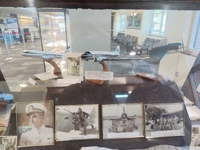 The image displays an aviation historical exhibit housed within a glass display case inside an airport terminal in the city of Park Hill, United States. The display case contains three model airplanes and four framed photographs. From left to right, the model aircraft include a white commercial passenger jet with green stripes, a white multi-engine propeller airliner with dark accents, and a dark military propeller aircraft marked with the number '61' on its tail and wing. All models are mounted on wooden stands. Small informational plaques with text are situated near the model bases. Below the models, resting on a dark surface, are four framed photographs. The leftmost is a color portrait of a man in a white naval officer's uniform. The subsequent three are black and white photographs: one depicts an individual in flight gear next to a military aircraft marked '35'; another shows a military jet aircraft viewed from the front with ground crew; and the last features a group of uniformed personnel posing in front of a military jet. Reflections on the display glass reveal parts of the surrounding airport environment, including a 'TSA Preโยฎ' sign and queue barriers on the left, and rows of grey airport seating, large windows, and framed artwork on the right.