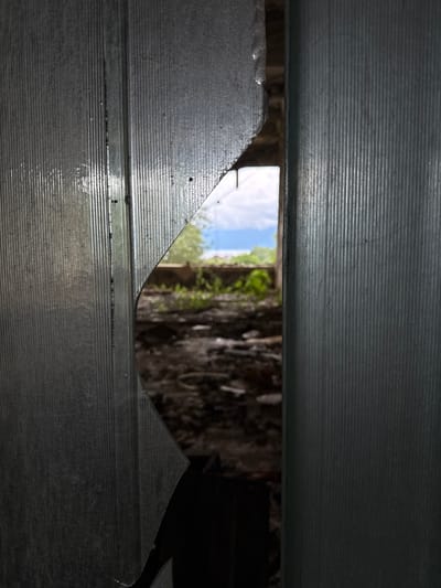 The image shows a view through a jagged hole in a corrugated translucent material, possibly polycarbonate, that appears to be part of a greenhouse or shed structure. The material is grey and shows signs of weathering and damage, with sharp, uneven edges around the opening. Through the hole, a blurry outdoor scene is visible. It depicts overgrown vegetation, debris on the ground, and a bright sky with white clouds. In the distance, hints of structures and greenery suggest an abandoned or neglected outdoor area. The overall impression is one of decay and disuse. The location is identified as Unknown, Abkhazia.