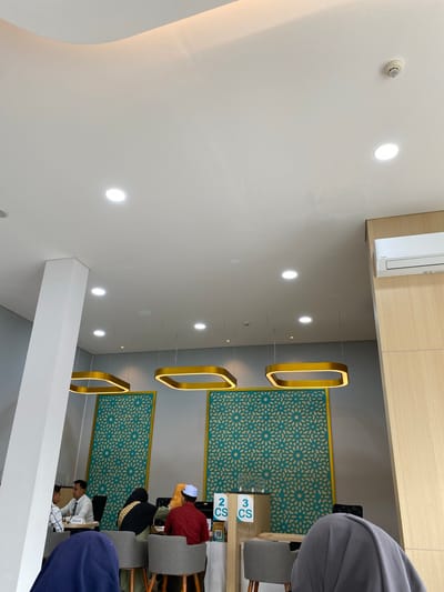 The image depicts the interior of what appears to be a government office or a service center in Lhoksukon, Indonesia. The scene is brightly lit with recessed ceiling lights and decorative golden pendant lights with a square-ish frame design. The walls are painted in a neutral gray color, with one section featuring an elaborate teal patterned screen. A light wood-paneled wall is visible on the right, housing a white air conditioning unit.
In the foreground, a white pillar is present on the left side. Further into the room, several individuals are seated. On the left, two men are engaged in conversation at a table; one man is wearing a white shirt with the name tag "FADLAN" visible. Behind them, two women are seated, one wearing a black hijab with a gold pattern and the other a yellow hijab. A man wearing a red shirt and a white cap is seated at a counter on the right. Two signs displaying "2 CS" and "3 CS" are visible on the counter. Several gray chairs are arranged around tables and counters. The overall atmosphere suggests an orderly administrative or customer service environment.