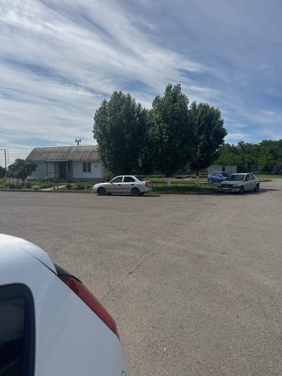 In Shakhty City District, Russia, this image captures an outdoor scene on a paved surface, likely a road or parking lot, under a sky with scattered clouds. In the background, a light blue house with a corrugated metal roof is visible, fronted by a small garden area with flowering bushes and some landscaping. Two large, mature trees with dense green foliage stand prominently near the house, their branches creating a broad canopy. Several cars are parked in the vicinity: a silver sedan is parked in front of the house, and further down, a silver sedan and a blue hatchback are visible. The foreground is dominated by the asphalt surface and the partial view of a white car from a low, rear-facing perspective. The overall impression is that of a quiet, residential or semi-rural area.