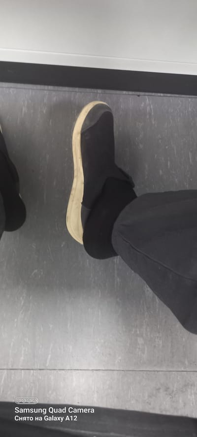 The image shows a close-up of a person's feet wearing black slip-on shoes with a light-colored sole. The shoes appear to be made of a soft, flexible material, possibly neoprene or a similar fabric, and have a rubberized toe cap. Black socks are visible peeking out from the top of the shoes. The person is wearing dark pants. The scene is set on a gray, textured floor that appears to be linoleum or tile, possibly in a domestic or commercial indoor setting. The lighting is somewhat dim, suggesting it might be indoors with artificial lighting or taken at night.
In the lower right corner of the image, there is visible text in white, which appears to be a camera watermark. It reads: "Samsung Quad Camera" and below that, "Снято на Galaxy A12". This indicates the photo was taken with a Samsung Galaxy A12 phone.
There is no clear indication of the time of day or weather conditions. The focus is entirely on the footwear and lower legs of the person, with no other subjects or significant background details visible. The overall impression is a simple, utilitarian shot focused on the shoes. Given the location context provided as Levakant, Tajikistan, the scene could be anywhere within that region, but without further visual cues, a specific location within Levakant cannot be determined. The image is devoid of any emotional cues.