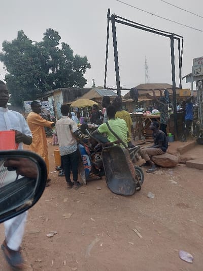A street scene shows several people, likely vendors or customers, clustered around a market stall. A wheelbarrow rests nearby. Above, a large metal frame suggests a signpost or structure. The scene is viewed from a vehicle, with a side mirror visible in the foreground, reflecting a hand and part of the arm. The ground appears to be dirt or reddish soil. The overall image suggests a bustling market atmosphere in a somewhat dusty or rural setting.