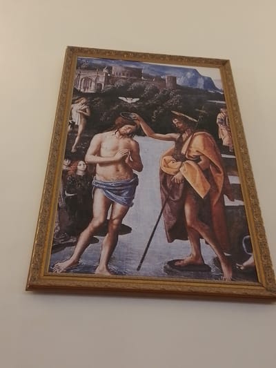 A framed print hangs on a white wall. The image is a depiction of the baptism of Christ. In the center of the painting, a pale-skinned, nude man wearing a blue cloth around his waist stands in water, while a darker-skinned, similarly dressed man pours water over his head. A dove hovers above the first man. To the left of the first man, two figures stand on the shore. Behind the men in the water, two other figures are partially visible on the shore. In the background, a landscape with buildings and mountains is depicted. The frame is ornate and gold-colored. The location is not within San Lorenzo, Paraguay.