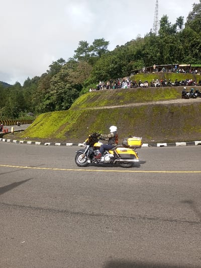 A yellow and black touring motorcycle, operated by a person wearing a white full-face helmet, a patterned jacket, dark trousers, and brown boots, is in motion on a curved asphalt road in Sandamal Eliya, Sri Lanka. The road features a solid yellow centerline and is bordered by a black and white striped curb on the left. To the right, a steep, moss-covered hillside rises, topped by a flat viewing area. Approximately 30-40 individuals are gathered on this upper level, observing the road below. Many of these individuals appear to be motorcyclists, with helmets visible and at least 15-20 additional motorcycles parked behind them. The background consists of dense green foliage, tall trees covering the hillsides, and a telecommunications tower positioned high on the ridge against a cloudy sky.