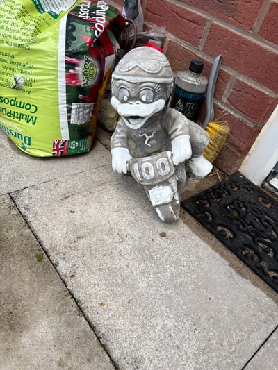 The image shows a corner of a property, seemingly in a domestic setting. A red brick wall runs along the left side of the frame, adjacent to a concrete walkway. A black, ornate doormat lies partially visible at the top. A grey, stone-like garden gnome figure is lying on its side on the walkway. Next to the gnome is a green and red bag of Durston Multi-Purpose Compost with the UK flag visible. A bottle of Auto Extreme detailing product, a grey handled brush, and a few other items are scattered nearby. The overall environment appears cluttered but domestic. It is not possible to place this image in Minsterley, UK.