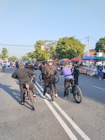 The image depicts a crowded street scene in Sragen, Indonesia, likely during a community event such as a Car Free Day or weekend market. The paved asphalt road, marked with distinct double white lines, is occupied by numerous individuals, including cyclists and pedestrians.
In the foreground, several individuals are positioned on bicycles, predominantly facing away from the camera. The closest cyclist on the left, viewed from the rear, wears a black long-sleeved shirt and black pants, riding a red bicycle. To their right, another cyclist in black sportswear and a helmet is on a black bicycle. Further right, a third cyclist wears a purple shirt, black shorts, and a helmet, also on a black bicycle. Other cyclists in similar attire are visible further into the midground. The presence of shadows stretching across the road towards the left suggests a morning or late afternoon setting.
The right side of the road is lined with an active market featuring numerous stalls, each sheltered by colorful umbrellas in shades of red, blue, and green. Various goods, including food items with visible signage such as "Sempul Goreng" and "Good Day," are displayed. Vendors attend to the stalls, and a small child in a blue shirt is visible near one of them. Beyond the market, traditional-style buildings, including a distinctive white gate structure, are partially visible.
The left side of the street and the background are densely lined with mature green trees, contributing to the open-air environment. Utility poles and overhead power lines are visible against the clear, light blue sky. In the midground, beyond the foreground cyclists, the road is filled with a large group of pedestrians, some walking casually, others appearing to browse the market. The overall scene conveys a bustling, communal atmosphere under bright natural light.