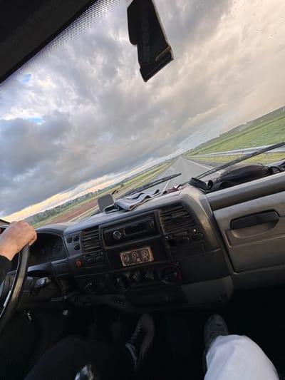 The image is a first-person perspective from inside a vehicle, likely a van or truck, driving on a highway. The driver's hands are visible on the steering wheel, and the dashboard with various controls and a stereo system is in the foreground. Outside the windshield, a rural landscape with green fields and a cloudy sky is visible. There are a few raindrops on the windshield. A car freshener hangs from the rearview mirror. The scene appears to be set in or around Bol'shaya Sosnova, Russia, given the context.