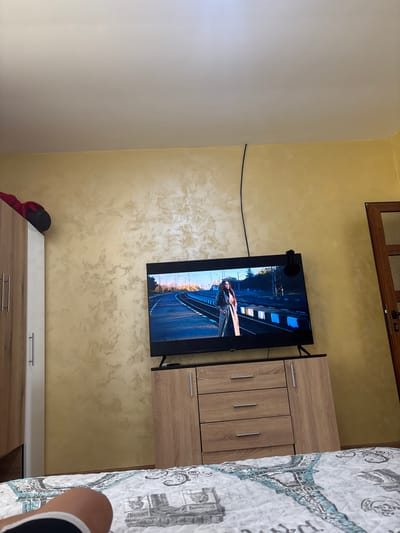 The image shows a portion of a room with a yellow-textured wall, a television mounted on it, a wardrobe or cabinet to the right of the television, and a bed partially visible in the right corner. On the television screen, an image of a woman lying on train tracks is displayed. Below the television, there's another low cabinet, and beside it, a black hat and a red piece of clothing are situated. The image does not provide enough information to confirm that it was taken in Septemvritsi, Bulgaria.