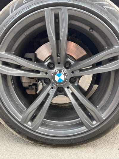 The media file is a close-up, high-angle shot of a BMW wheel and tire mounted on a vehicle. The wheel is a dark gray, multi-spoke alloy design with a BMW logo at the center. The tire is black with visible text on its sidewall, indicating its specifications: "245/45ZR19 102Y EXTRA LOAD," along with other markings related to tread composition and load capacity. The brake rotor and caliper are also visible behind the wheel spokes. The setting appears to be outdoors, with the wheel resting on a textured gray surface that resembles concrete or asphalt. The lighting suggests it is daytime. No people or specific events are depicted, only the automotive component.
