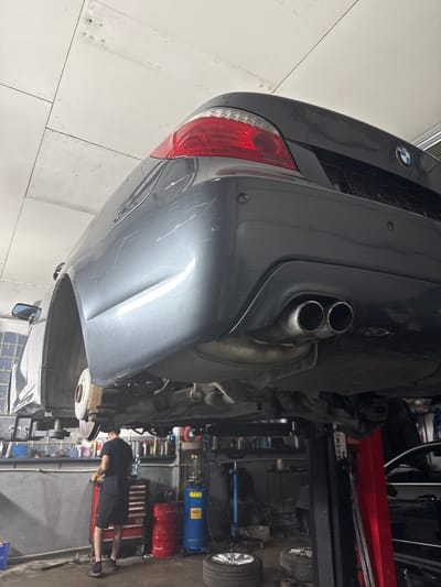 A person is working on the underside of a grey car that is on a lift in a garage. The car's rear bumper and exhaust pipes are visible. The person is wearing a black t-shirt and shorts and is bent over the car, appearing to be working on the brakes or suspension. The garage is filled with tools and equipment, including a red tool chest and a blue air compressor. The floor is concrete and there is a white tiled wall in the background. The lighting suggests it is daytime.