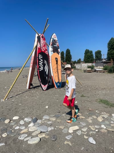 A young boy in a white t-shirt and red shorts stands on a pebble beach next to three paddleboards leaning against bamboo poles. The paddleboards are decorated with various designs, including geometric patterns and an astronaut. The beach is sandy with scattered rocks. In the background, there are trees, buildings, and a clear blue sky. The sea is visible in the distance, with a few people and a boat on the shore. The location appears to be Giachrypsh, Abkhazia, as suggested by the "MYCOP" graffiti visible on a wall in the background, a common sight in the region referring to the local waste management company.