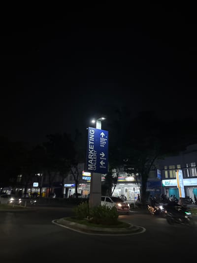 This is a nighttime photograph taken in South Tangerang, Indonesia. The image is dominated by darkness, with only illuminated elements providing visibility.
The primary subject is a blue directional signpost. It displays white text and arrows. One sign reads "MARKETING GALLERY" vertically, with an arrow pointing downwards. Another sign has two sections: the left side shows an arrow pointing left and text that appears to read "Fortune District". Below that, it says "Pinang" and "GAS". The right side of this sign shows an arrow pointing right with text "Venice" and "Verina".
In the background, there are several buildings with lit windows and illuminated signs, suggesting commercial establishments. One prominent sign appears to be for "BSI". There are also parked motorcycles and a white car visible, with their headlights on. The scene suggests a street or parking area in a commercial district at night. The lighting is artificial, coming from the signs, buildings, and vehicles.
The overall impression is of a busy urban environment at night, focused on navigation and commercial activity. The darkness emphasizes the illuminated signs and vehicles, creating a contrast.