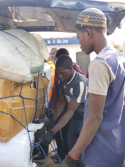 The scene depicts an outdoor cargo loading operation in Unguwa Uku Kauyen Alu, Nigeria. Four adult males are visible around the open rear cargo area of a dark-colored vehicle, likely an SUV or minivan. In the immediate foreground to the right, a man wearing a dark blue and light-colored short-sleeved top and a black and gold kufi cap stands in profile, observing the activity. His left arm shows a light dusting of dirt. To his left, a man in a dark grey short-sleeved athletic shirt with white stripes on the sleeves is actively engaged in tying or adjusting a blue rope net around numerous white sacks and cardboard boxes packed into the vehicle's trunk. He wears black work gloves with red wristbands, and his arms are also covered in a light layer of dust. Behind him, another man in a dark red long-sleeved shirt is partially obscured. Further back, a bearded man in an orange and white striped shirt is visible, looking towards the camera with a slight smile. The cargo includes several large white woven sacks and multiple brown cardboard boxes, secured by the blue rope net. In the background, a building with a blue awning or sign displaying "Eat & Take Away" in white text is visible, along with a grey door on an adjacent structure. The lighting suggests daytime conditions.
