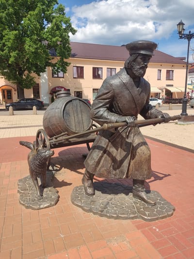 The image presents a bronze sculpture prominently situated on a public square in Staraya Russa, Russia, under a bright blue sky with scattered white cumulus clouds. The central sculpture depicts a bearded man, dressed in a long coat and a peaked cap, actively pulling a two-wheeled cart. The man is rendered in a crouched, forward-leaning posture, gripping the cart's handles with both hands. The cart carries a large, horizontally positioned wooden barrel, detailed with staves and metal hoops, and a prominent bung on its upper surface. Accompanying the man, to his left and slightly ahead, is a bronze figure of a wolf or dog-like animal, characterized by distinct ears and small, wing-like formations on its back, looking forward and slightly upward.

The sculpture, executed in a dark bronze patina, rests on irregularly shaped bronze bases that transition into the surrounding ground. The ground consists of red brick pavers laid in a running bond pattern, illuminated by direct sunlight, casting clear shadows from the sculpture. In the background, a light peach-colored, two-story building with a dark brown tiled roof is visible, featuring multiple rectangular windows with dark frames. A red sign with white Cyrillic lettering, indicative of a cafe, is affixed to the building's facade. To the left, a large deciduous tree with dense green foliage partially obscures the building. On the right, a black, ornate lamppost with two lanterns stands upright. Beyond the primary building, a paved street is visible, with a dark sedan parked on the left and a white vehicle further in the distance. The overall environment suggests a well-maintained urban area.