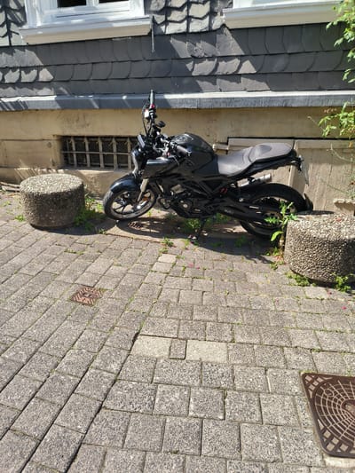 The image shows a black motorcycle parked on a cobblestone sidewalk in front of a building. The building has a facade with grey slate tiles and a white-trimmed window. Below the window, there is a metal grate covering a small opening. Two cylindrical concrete bollards are placed on the sidewalk, one on each side of the motorcycle. Green plants and a few fallen pink petals are scattered on the cobblestones. The scene appears to be set outdoors during the daytime, with sunlight casting shadows. The ground features a manhole cover with the word "GAS" visible on it. The overall setting suggests an urban environment in Wuppertal, Germany, likely a residential or commercial street. No people are visible in the image, and the motorcycle is stationary. The image quality is clear, capturing the details of the motorcycle, the building's exterior, and the ground surface.