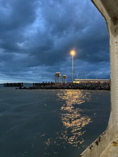This is a nighttime outdoor photograph of a pier or seawall at dusk. The sky is dark and cloudy, with dramatic, heavy clouds casting a somber mood. A single bright lamp post illuminates the scene, its light reflecting off the dark, choppy water. In the distance, there are structures that appear to be part of a dock or industrial area, including pilings and a long, low building with lights. The foreground shows the texture of the water and the reflection of the lamp. The right edge of the frame is partially occupied by a white, weathered railing or structure, possibly from a boat or a walkway. The overall impression is one of a utilitarian coastal environment at the end of the day, with a sense of impending weather or a transition in light.