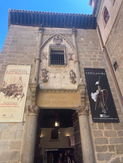 The image shows the entrance to the "Posada de la Hermandad" in Toledo, Spain. The building is constructed of stone, with a red-tiled roof. Two banners hang on either side of the entrance. The left banner advertises an exhibition of catapults and siege machines, and the right banner advertises an exhibition about the Templars. The entrance itself is a large stone archway with two large wooden doors. Above the archway, there is a small window with a wrought-iron grille. Two small stone statues are visible on either side of the window, and two larger statues are positioned on columns flanking the entrance. A wrought-iron lantern hangs inside the entrance. The sky is clear and blue. The scene is a street view of an historic building.