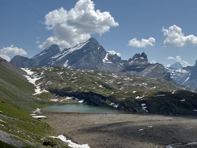 This is a wide, high-angle, daylight, landscape photograph of a mountainous alpine environment in Leukerbad, Switzerland. The scene is dominated by rugged, snow-capped mountain peaks under a blue sky with scattered white clouds. In the foreground, a steep, grassy slope descends towards a tranquil, turquoise lake. The lake is bordered by a rocky, barren shoreline and patches of snow. A thin cable, part of a cable car system, stretches diagonally across the upper portion of the frame, connecting to the distant mountains. The terrain is rugged and uneven, with visible rock formations and sparse vegetation. The overall impression is one of a remote and wild natural landscape.