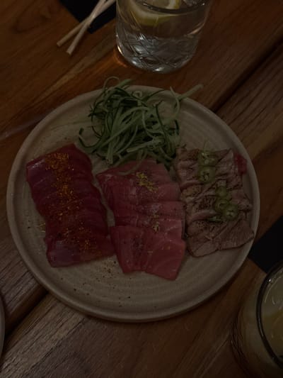 This image depicts a close-up overhead view of a meal served on a wooden table in a dimly lit setting. The central subject is a light-colored, round ceramic plate presenting three distinct preparations of tuna sashimi or tataki.

On the left side of the plate, there are multiple slices of deep red raw tuna, lightly sprinkled with a reddish spice blend. In the middle section, several slices of lighter pinkish-red raw tuna are arranged, topped with fine yellow zest, possibly lemon. The right portion of the plate features slices of seared tuna (tataki style), exhibiting distinct grill marks and garnished with thin green chili slices, likely jalapeño or serrano. Above these tuna preparations, a pile of thinly julienned green cucumber or similar vegetable is arranged.

To the upper right of the plate, a clear glass contains a beverage with ice and lemon slices. Two light-colored chopsticks rest on the wooden table surface above and to the left of the plate. A partial view of a second drink in a ribbed glass is visible in the lower right corner. The table surface is made of dark wood with a visible grain pattern, and the lighting is subdued, suggesting an indoor dining environment such as a restaurant or bar.

No specific visual identifiers within the image provide information about a precise location in the city of Guadalajara, Mexico.