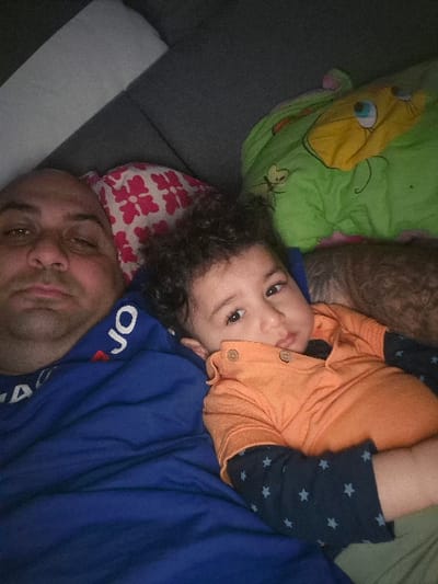 The scene depicts two individuals, an adult male and a child, lying on a dark grey upholstered surface within an indoor domestic setting in Melle, Belgium. The adult male, with a partially bald head and short facial hair, is positioned on his back, gazing directly at the camera. He is partially covered by a blue blanket and wears a blue shirt with white lettering. To his right, a toddler with dark, curly hair lies on their back, their head turned towards the camera with a direct gaze. The child wears an orange long-sleeved polo shirt with three buttons over a dark blue long-sleeved shirt patterned with light blue stars. A red and white patterned pillow is visible behind the adult male's head, and a green blanket featuring a yellow cartoon character is situated near the child's right side. The ambient lighting suggests an interior space.