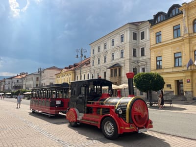 In Banská Bystrica, Slovakia, on a sunny day, a red and black tourist train is parked on a cobblestone street in front of historical buildings. The train consists of a locomotive with the inscription "NEUSCHWANSTEIN EXPRESS" on its side and several passenger carriages. A woman and a child are walking away from the train, while a man walks towards the train. The buildings flanking the street exhibit traditional European architecture with pastel-colored facades and ornate detailing. The sky is partly cloudy, with the sun casting shadows on the street and buildings.